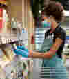 A woman wearing an apron and gloves stocks products on the shelves in a supermarket aisle.