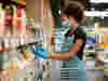 A woman wearing an apron and gloves stocks products on the shelves in a supermarket aisle.
