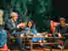 Four speakers have a discussion sitting on sofas, with a green forest-themed screen display behind.