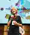 A woman wearing a lanyard and holding a microphone speaks, with a map of Europe on the projector screen in the background.